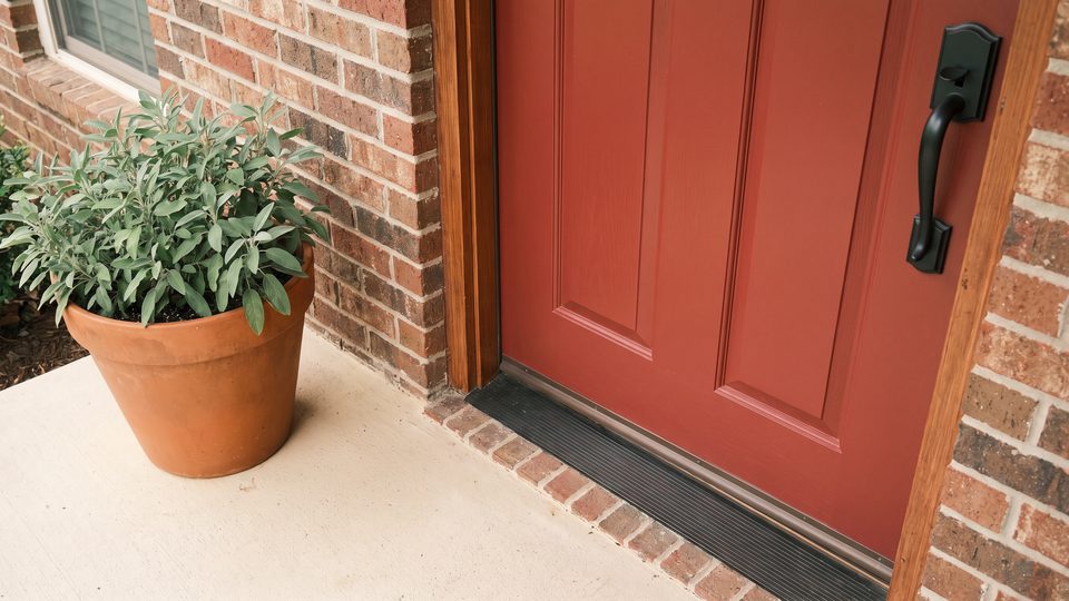 A brick-red front door with black handle hardware, a doormat and a potted sage plant on a concrete step