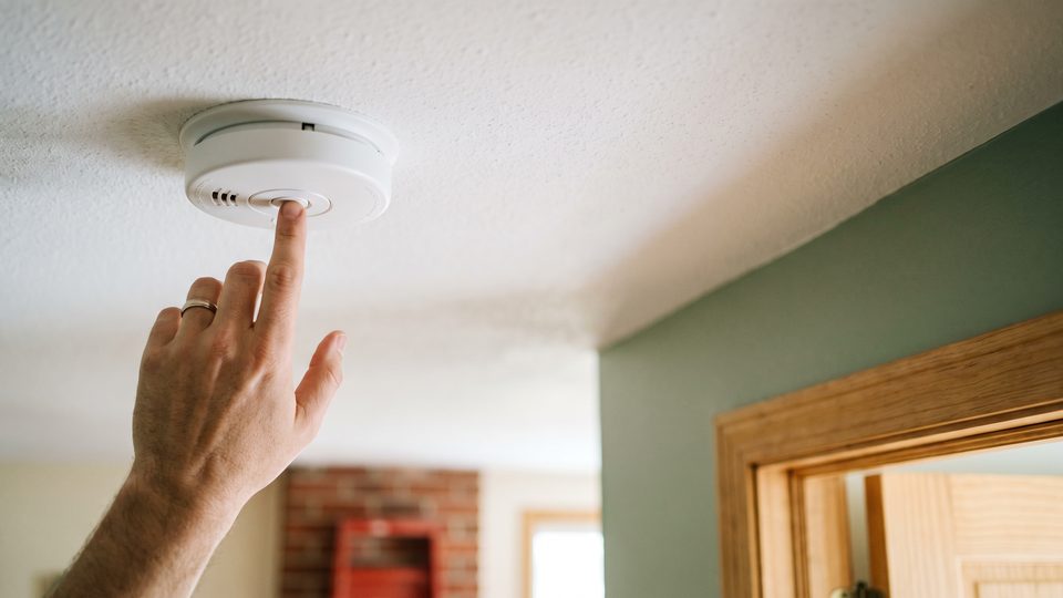 A finger presses the test button on a white ceiling smoke alarm in a hallway