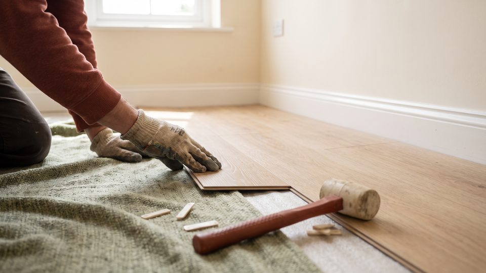 Gloved hands fit a new wood-look plank into a floor over an underlayment sheet, with a rubber mallet nearby